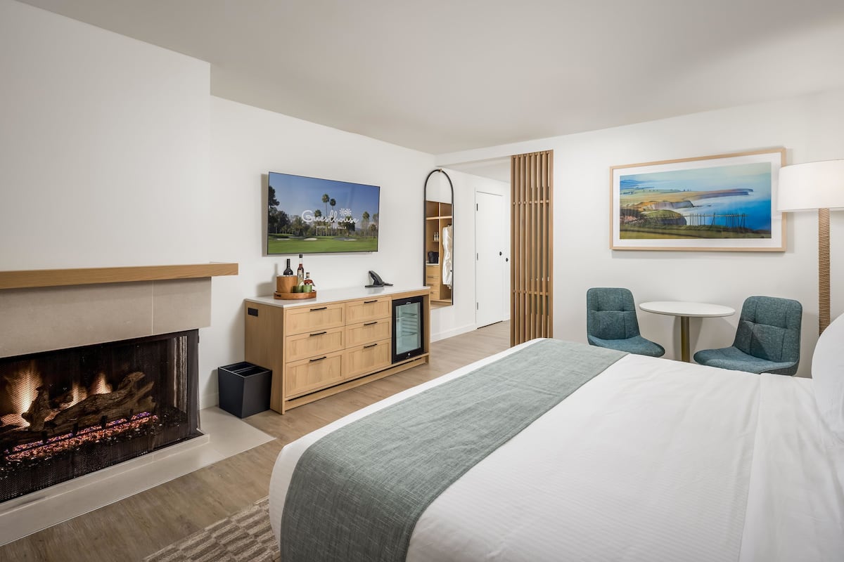 A hotel room with a fireplace, a bed, a desk, two chairs, a round table, and two paintings on the walls.