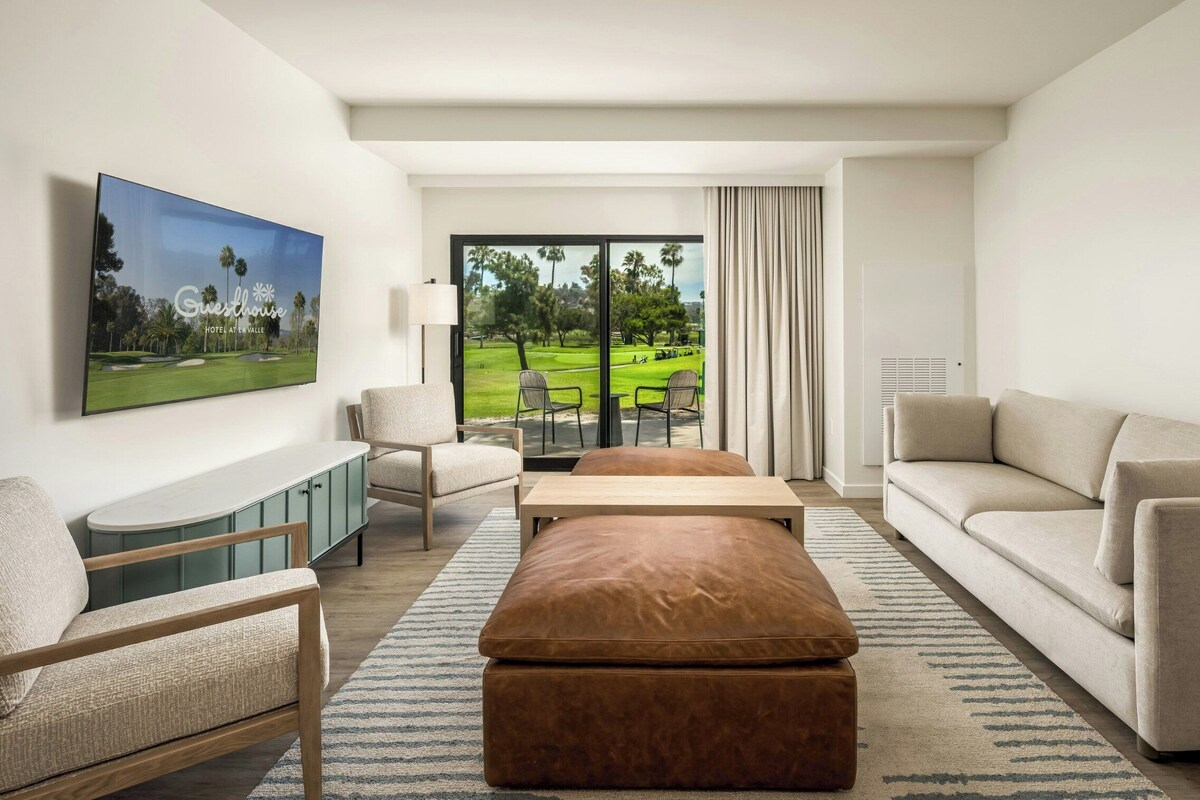 A modern hotel room with a large window offering a view of a golf course, a flat-screen TV mounted on the wall, a sofa, a chair, and a small table.