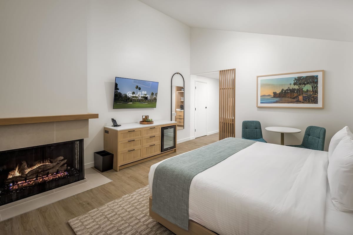 A modern hotel room with a fireplace, a bed, a desk with a chair, a small table, and artwork on the walls.