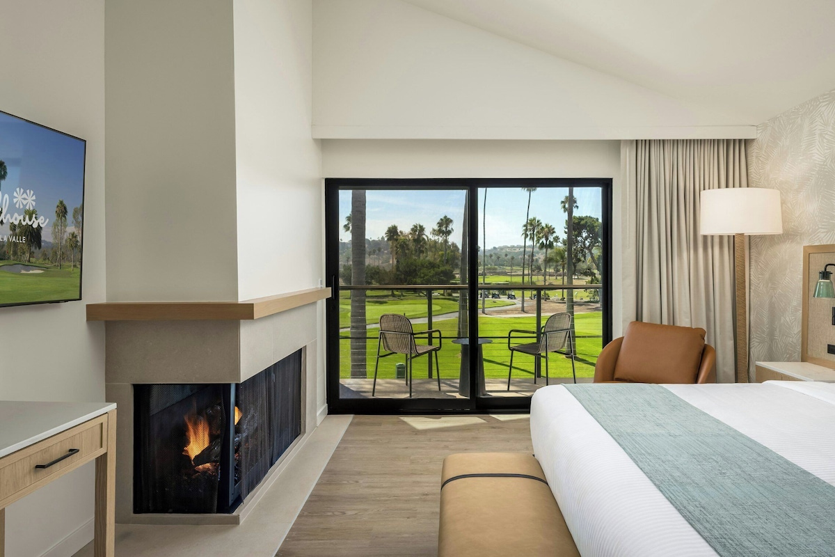 A hotel room with a large bed, a fireplace, a TV, and a balcony with a golf course view.