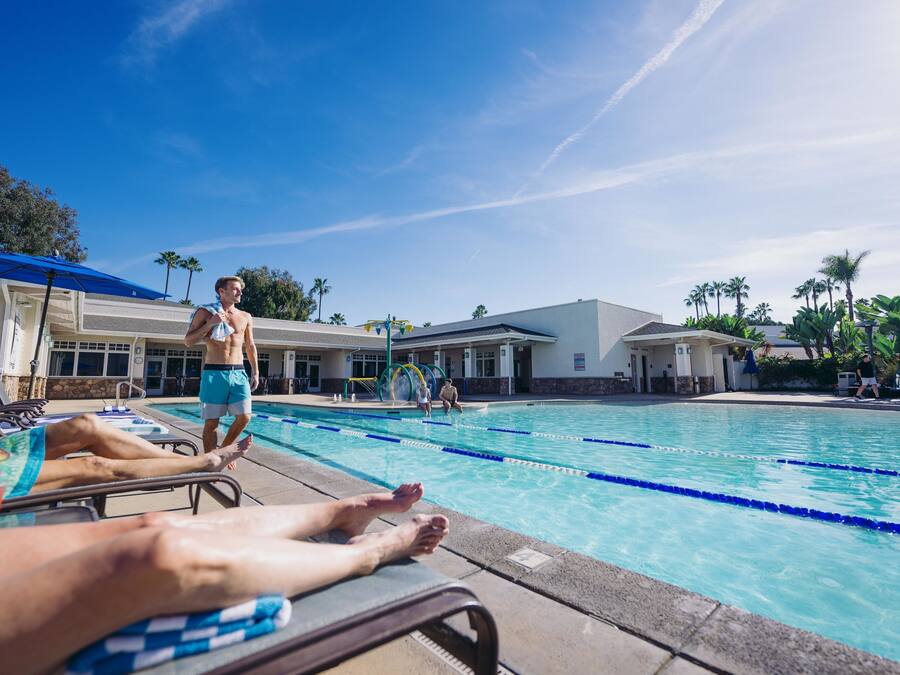 A swimming pool area with lounge chairs, a pool with lane dividers, and a building with a flat roof and windows.