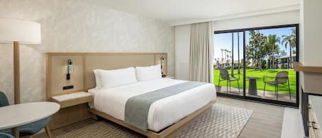 A hotel room with a bed, a desk, a chair, and a view of a green lawn and buildings.