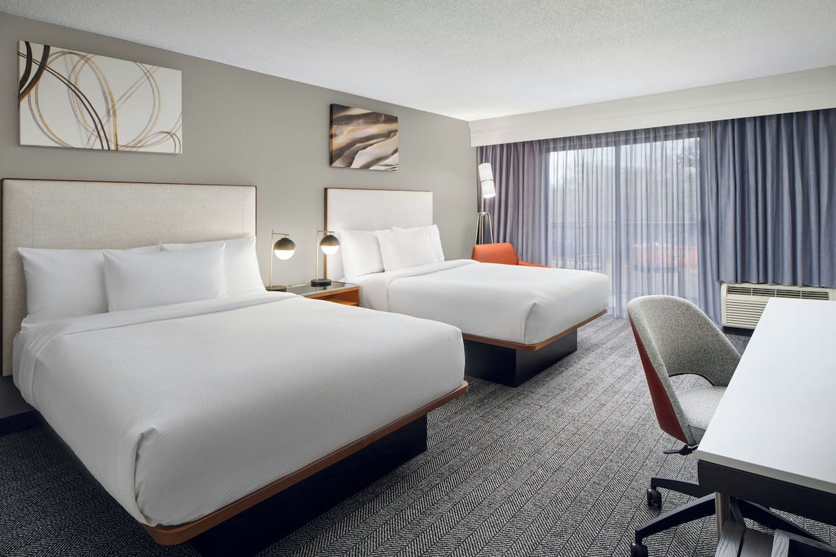 A hotel room with two beds, a desk, a chair, and a large window with curtains.