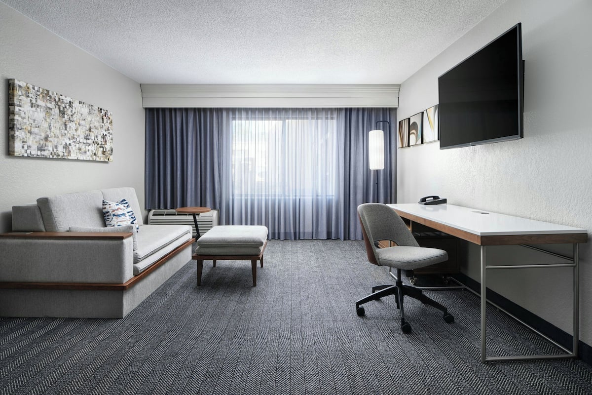 A modern hotel room with a sofa, a desk, a chair, a flat-screen TV, and a large window with curtains.