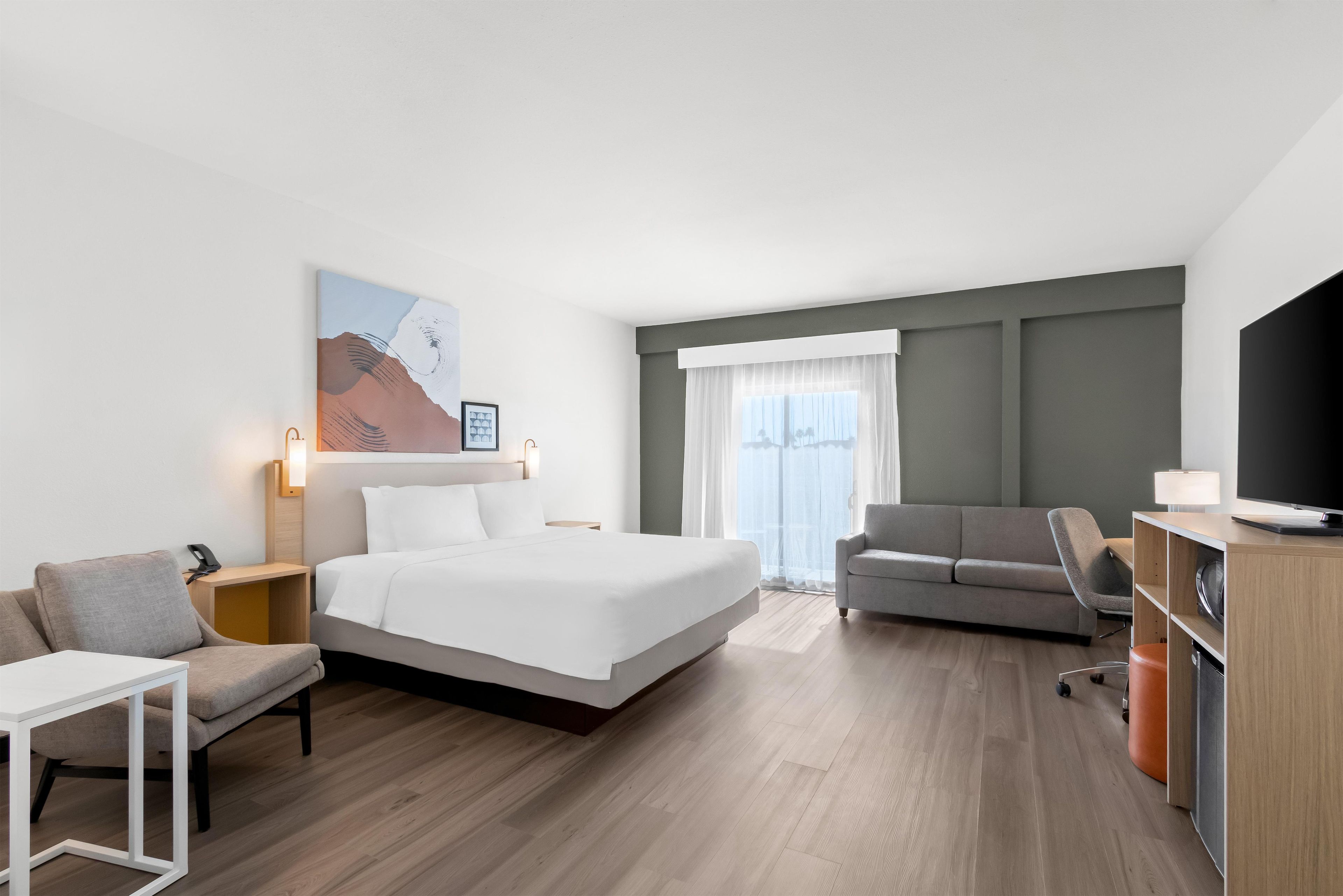 Habitación de hotel con cama, sofá, escritorio, televisor y un amplio ventanal con cortinas.