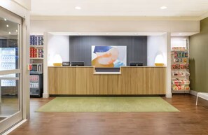 A reception area with a wooden counter, a large abstract painting above it, and a green carpet. There are shelves with bottled water and snacks on the right. - Spark by Hilton Fountain Hills Scottsdale (Fountain Hills)