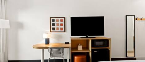 A hotel room with a bed, a desk with a chair, a television, a mirror, and a small cabinet.