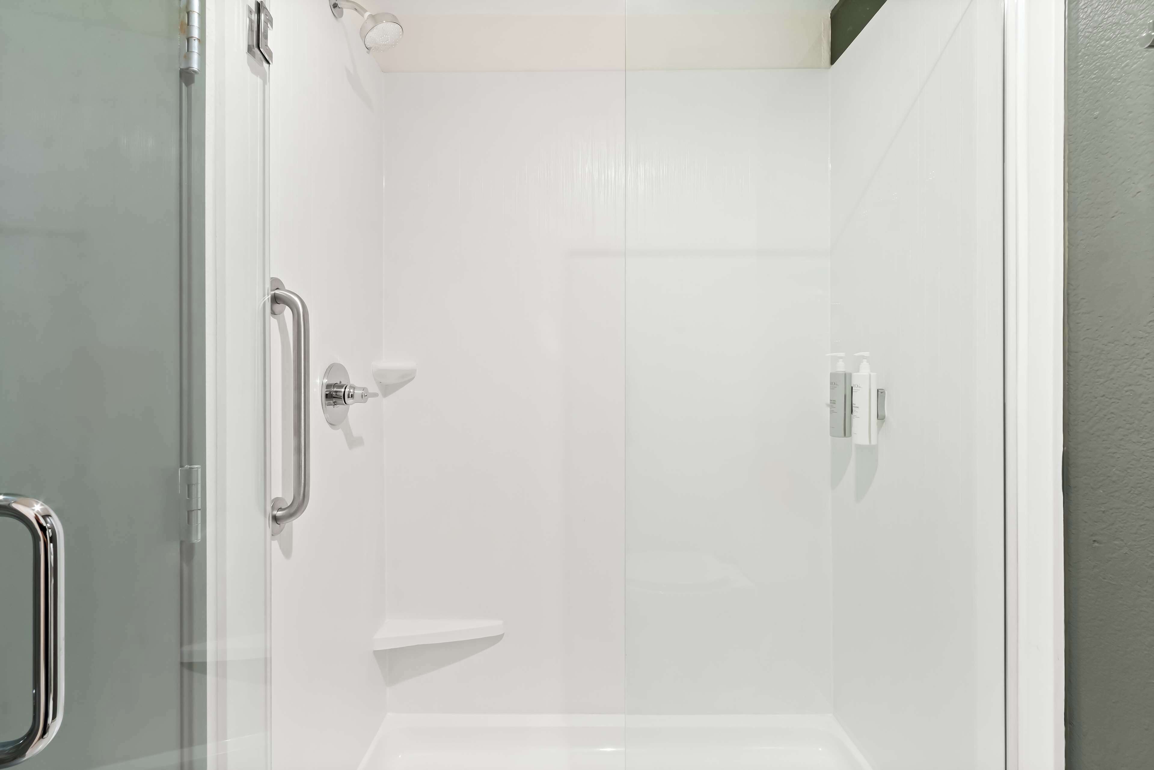 A glass-enclosed shower stall with a white tiled wall, a handheld showerhead, and a handheld shower control.