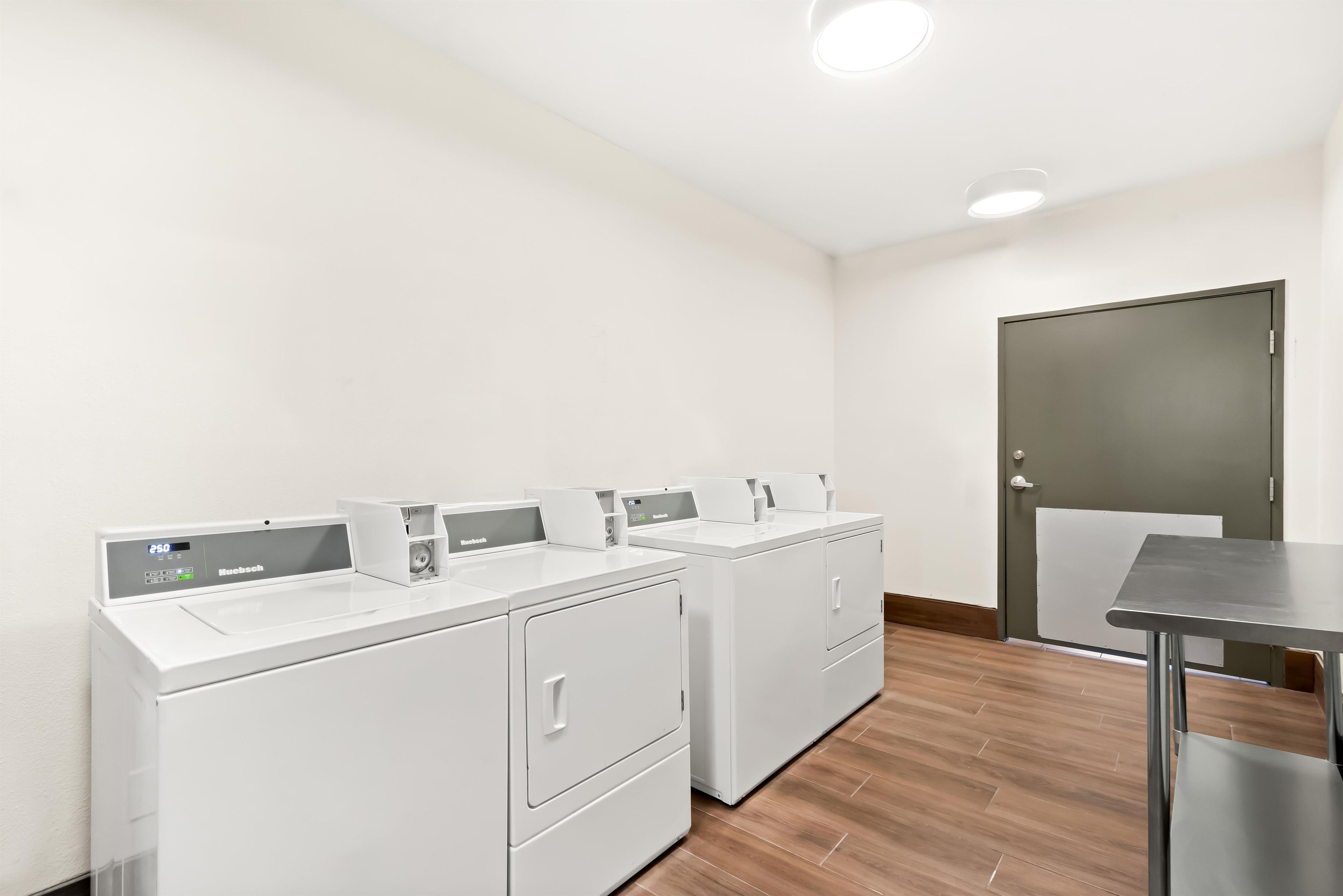 A laundry facility with four white washing machines, a grey door, and a small table.