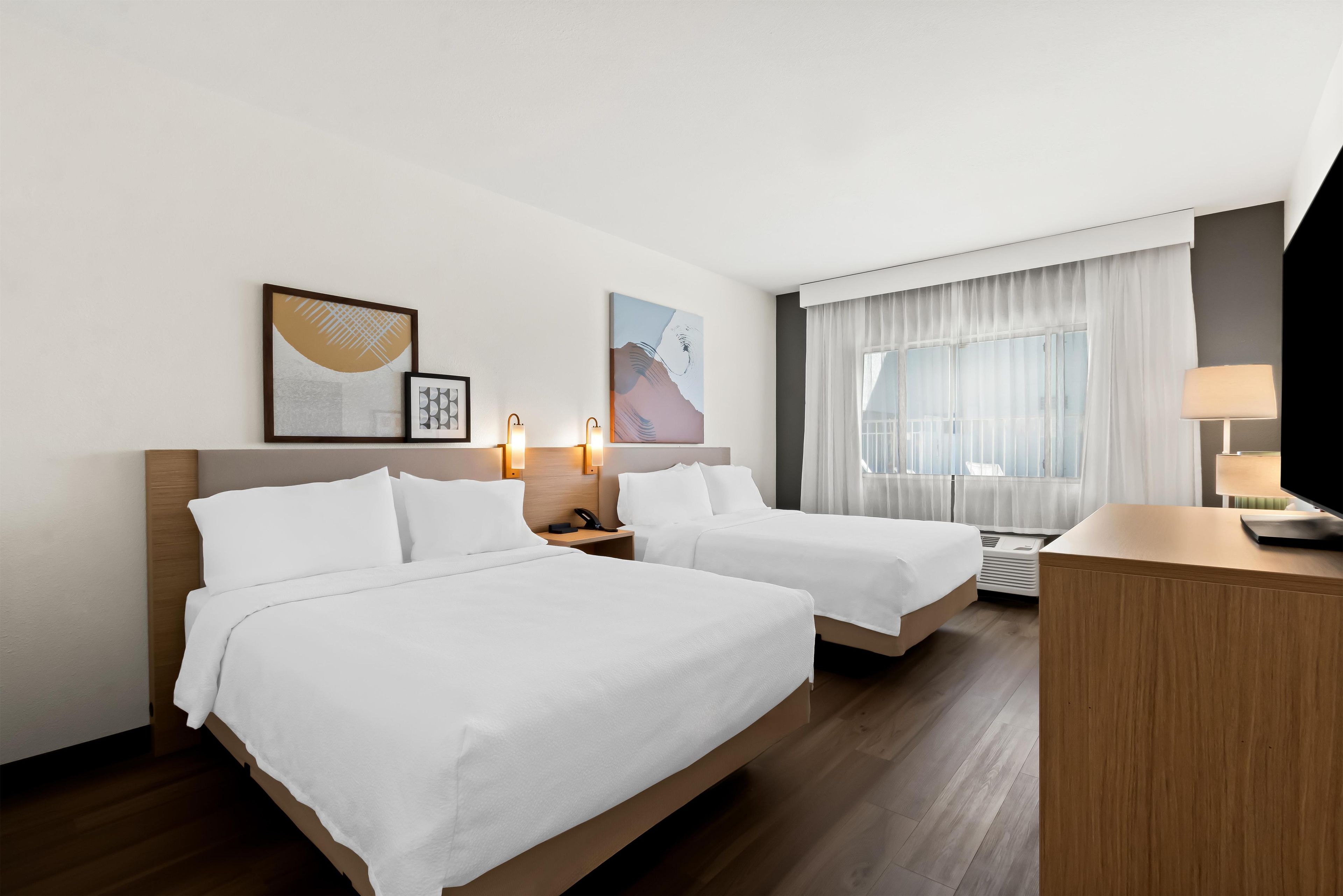 A hotel room with two beds, a desk, a lamp, and artwork on the wall.