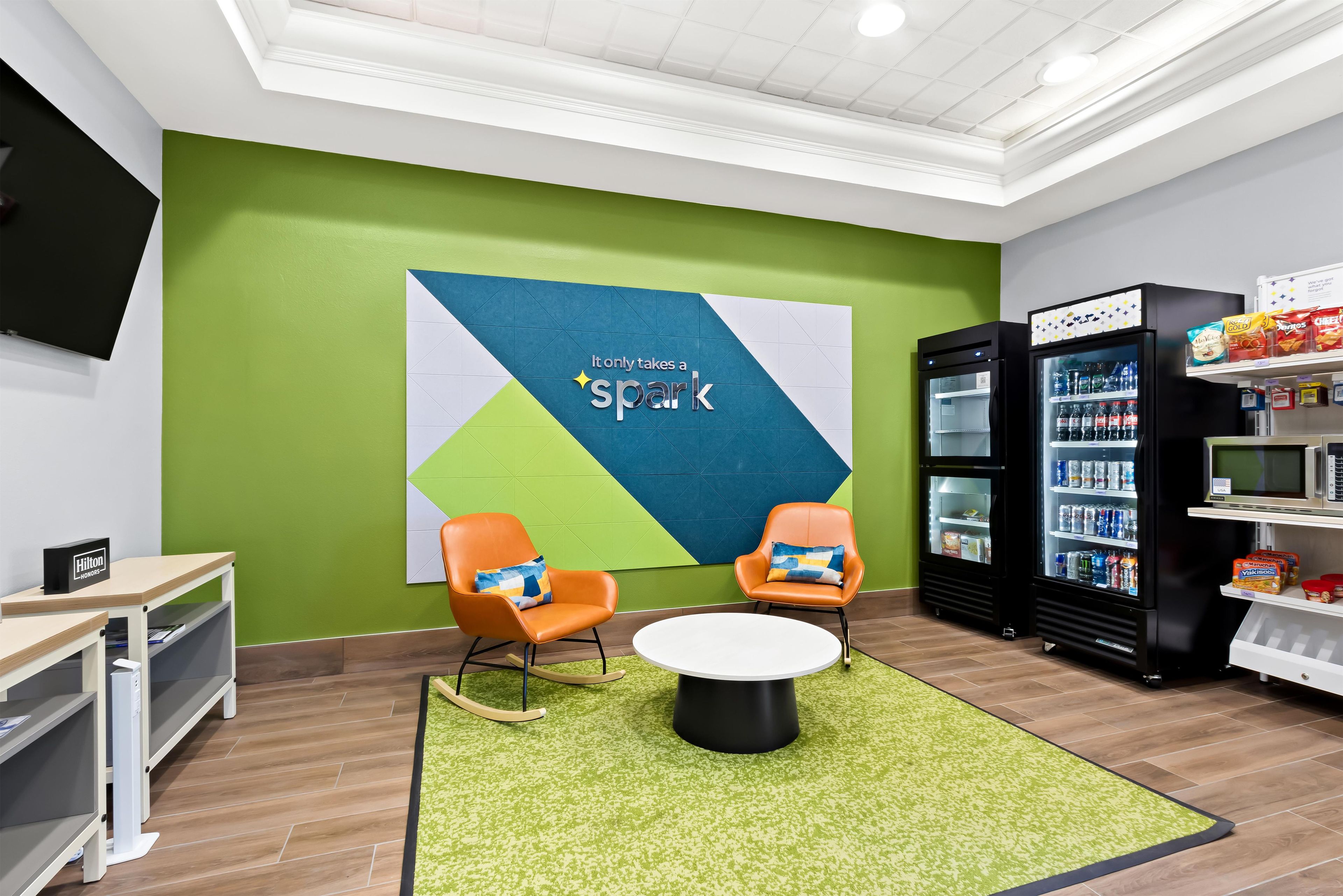 A modern lounge area with a green wall, two orange chairs, a white coffee table, and a vending machine.