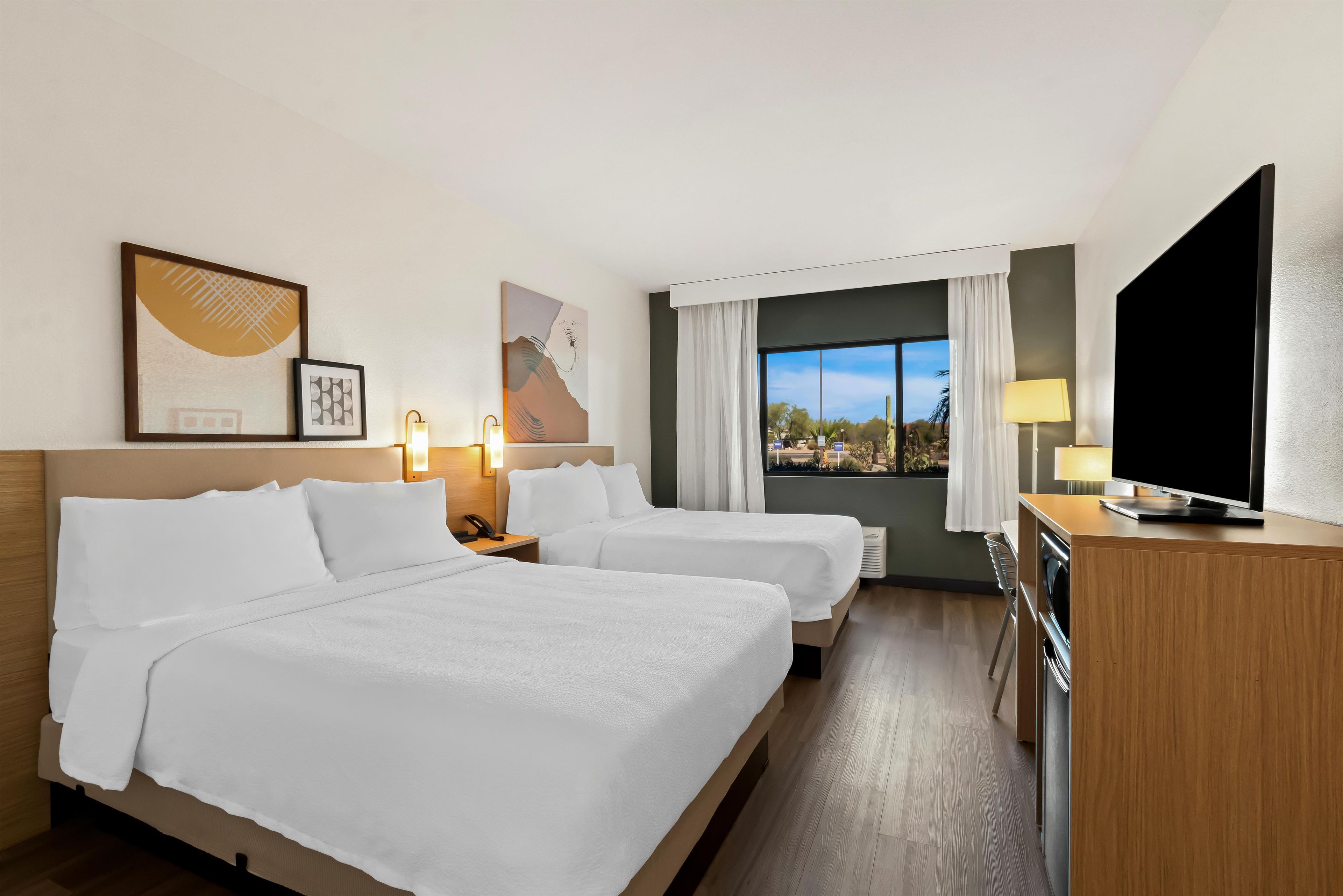 Habitación de hotel con dos camas, una televisión, un escritorio con lámpara y cuadros en la pared.