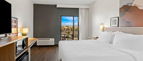 A hotel room with a bed, a TV, a desk with a lamp, a heater, and a window with a view.