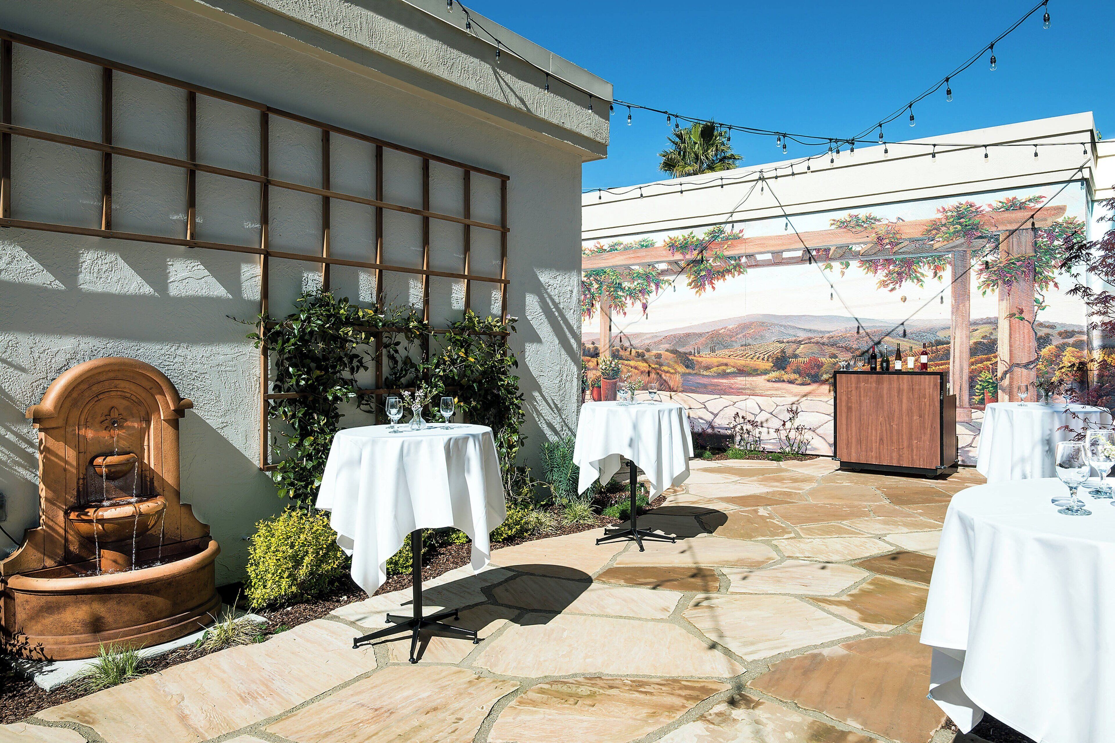 An outdoor patio with stone flooring, white tables, and a fountain.