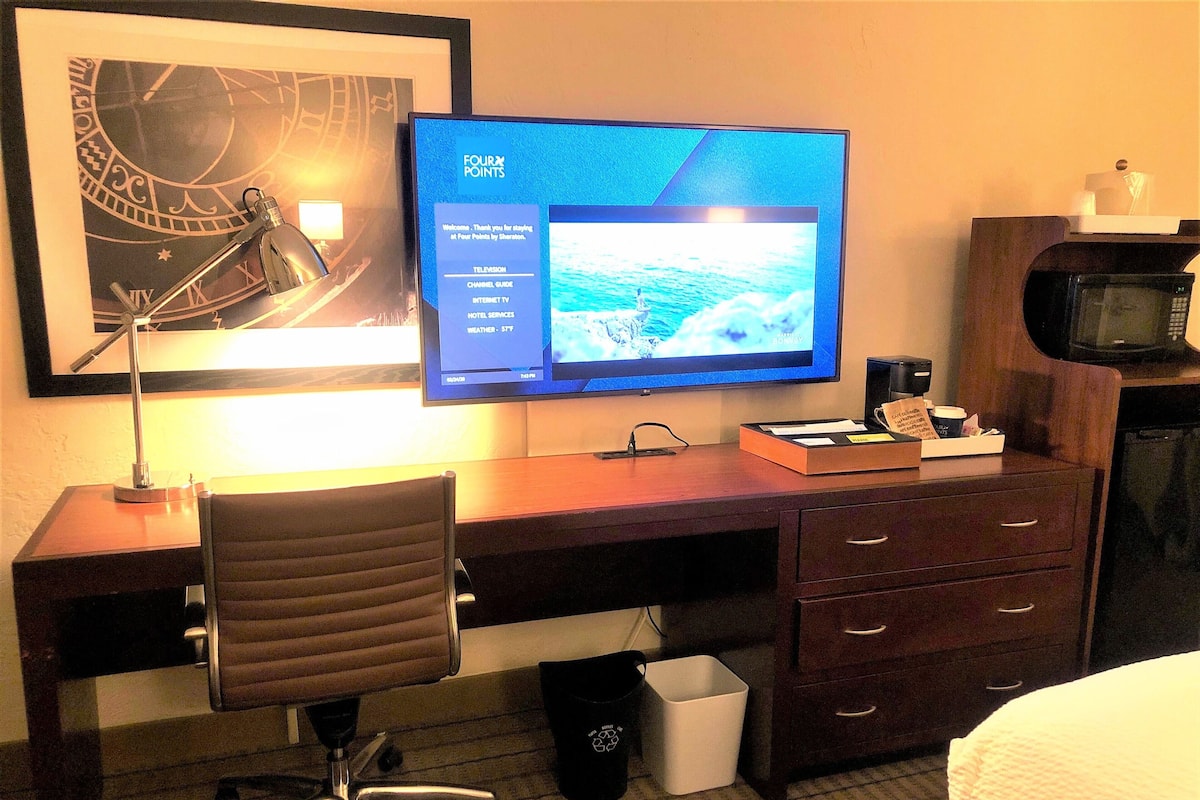 A hotel room with a desk, a chair, a television displaying a channel guide, a microwave, and a trash bin.