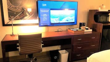 A hotel room with a desk, a chair, a television displaying a channel guide, a microwave, and a trash bin.