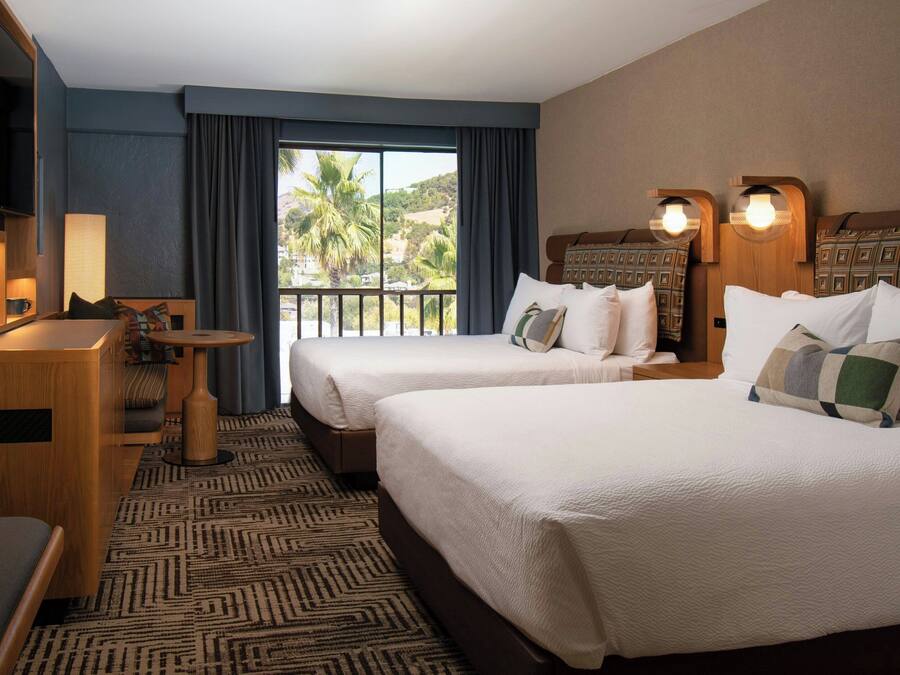 A hotel room with two beds, a desk, a chair, a lamp, and a large window with a view of palm trees.