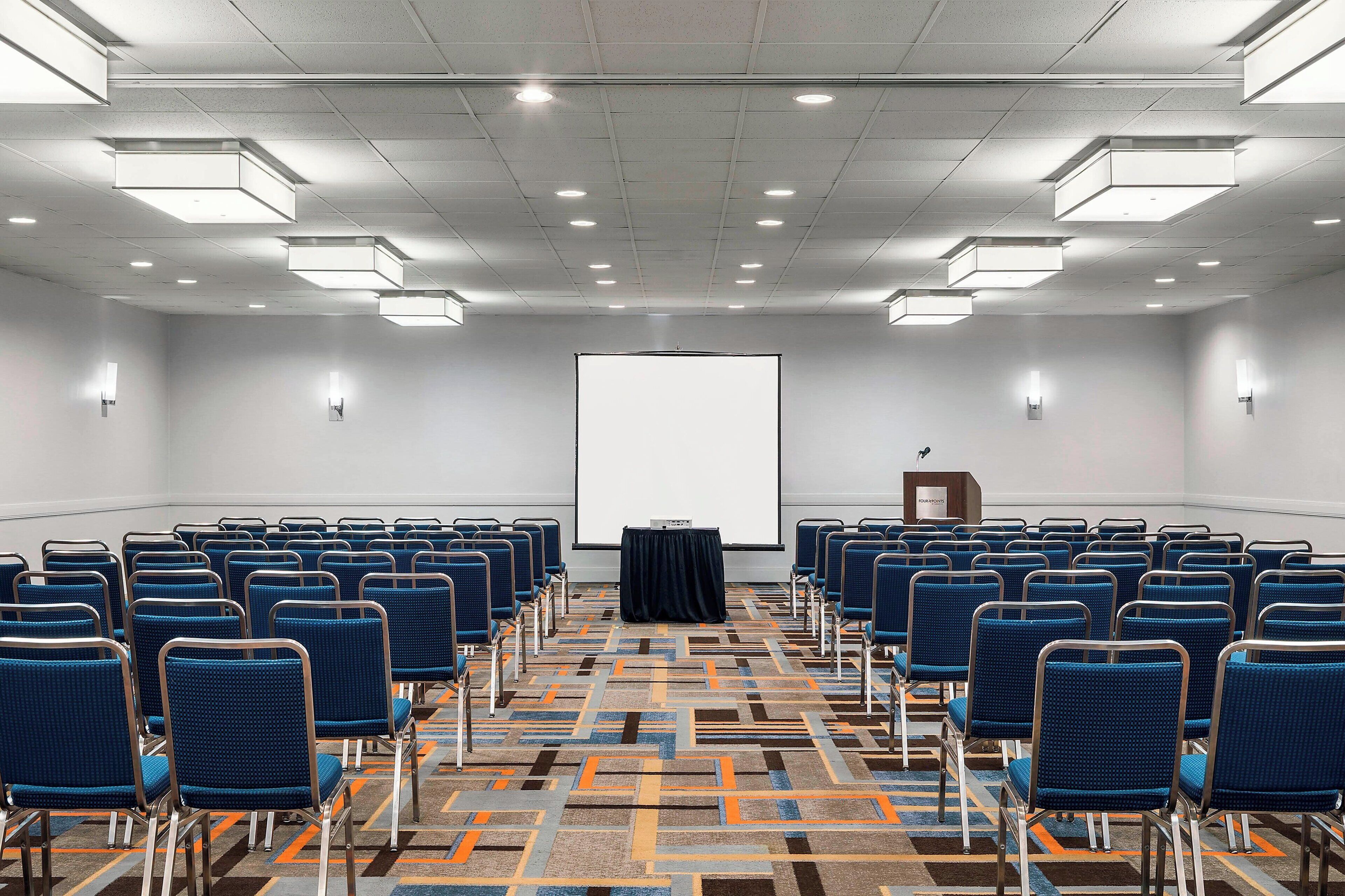 Un salón de conferencias con filas de sillas azules, una pantalla de proyector y un atril.