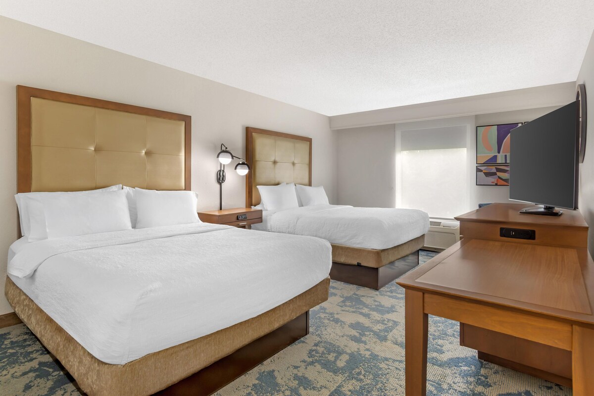 A hotel room with two beds, a desk, a TV, and a window.