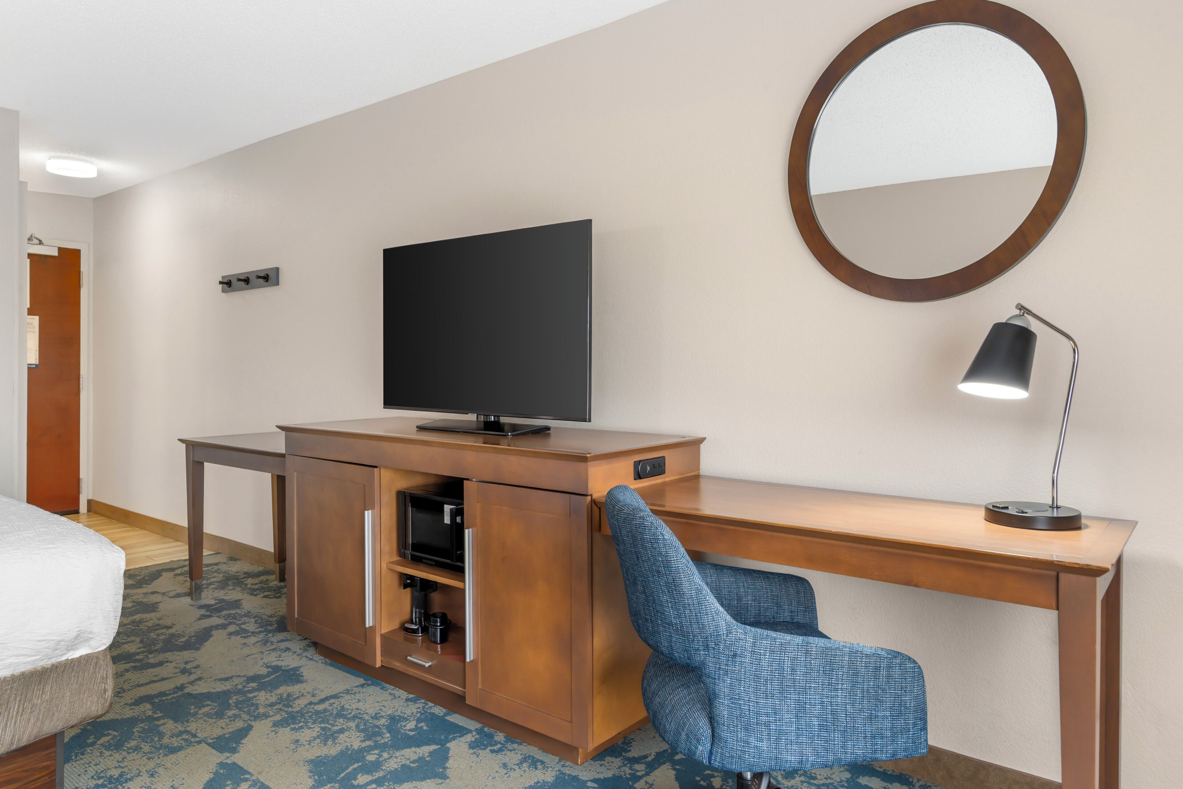 Habitación de hotel con cama, escritorio con televisor, silla, lámpara de escritorio y espejo en la pared.