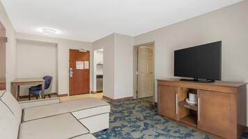 Habitación de hotel con sofá, televisor sobre un mueble de madera, escritorio con silla y puerta que da a un baño.