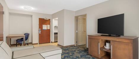 A hotel room with a sofa, a television on a wooden cabinet, a desk with a chair, and a door leading to a bathroom.