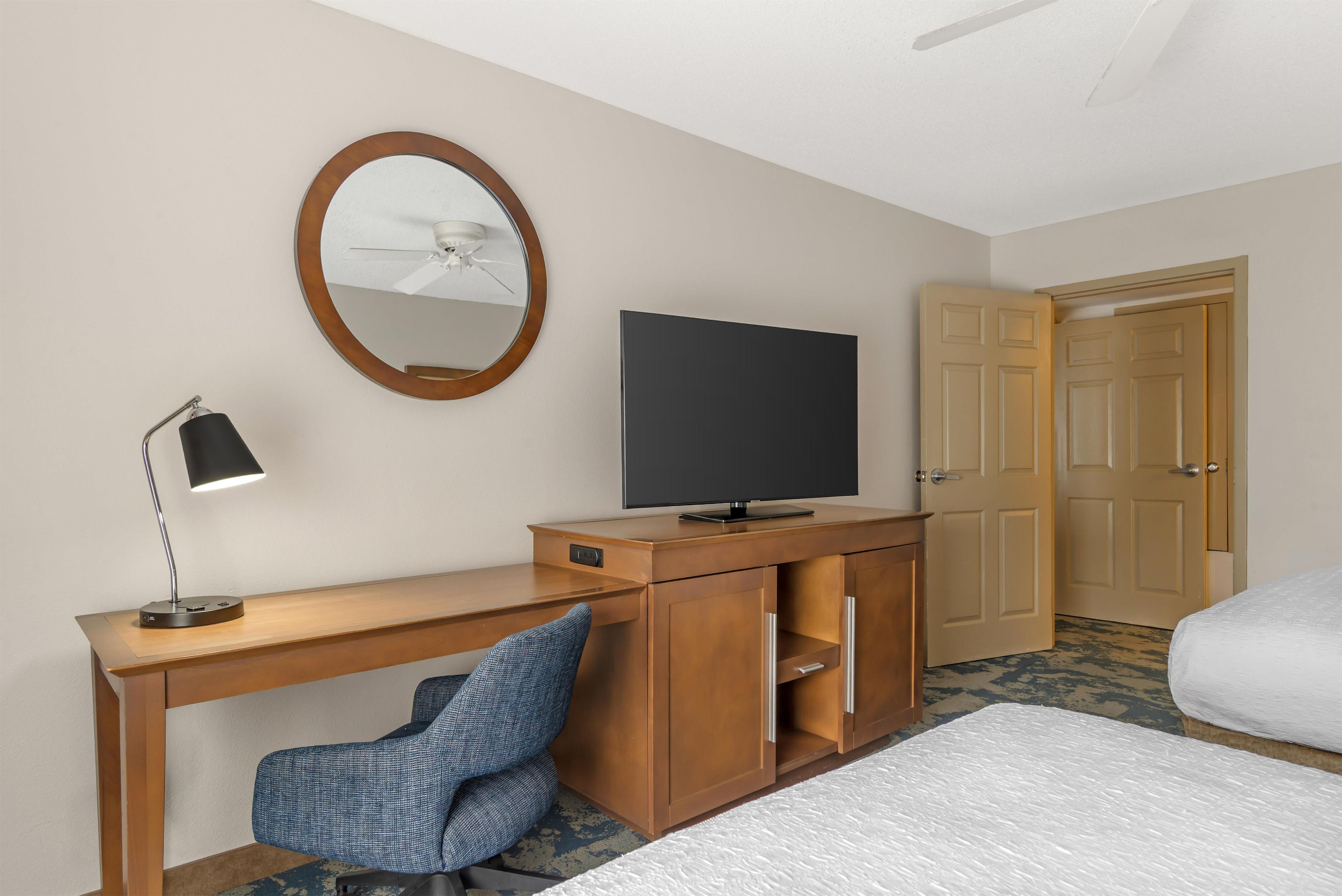 Een hotelkamer met een houten bureau, een televisie, een spiegel, een plafondventilator en een bed.