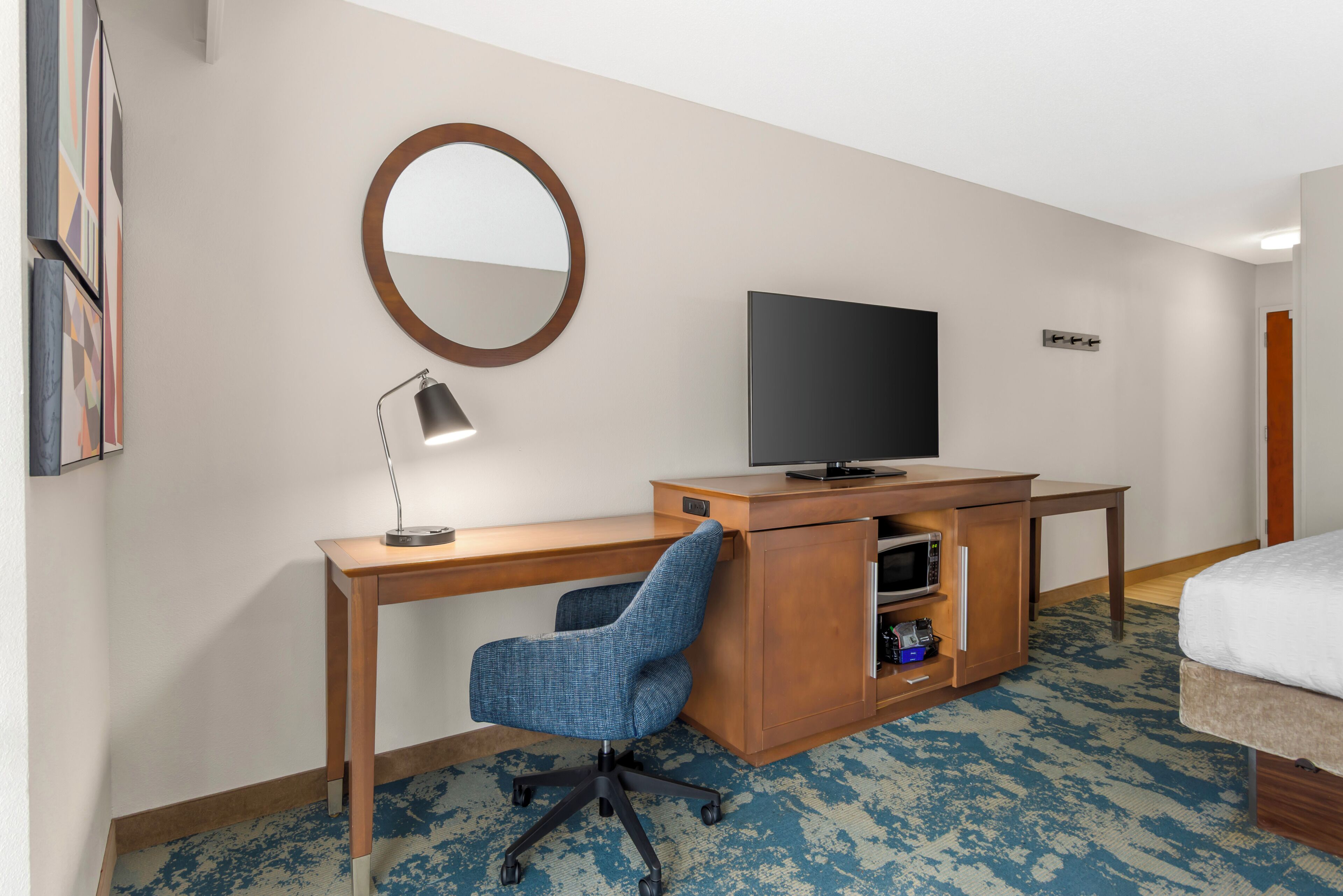 A hotel room with a wooden desk, a blue office chair, a television, a mirror, and a bed.
