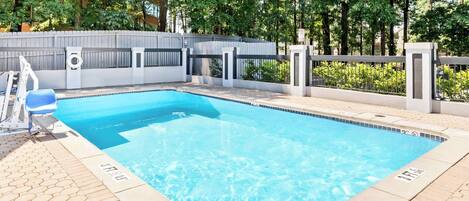 Una piscina rectangular con borde de azulejos, rodeada por un suelo empedrado y una valla blanca.
