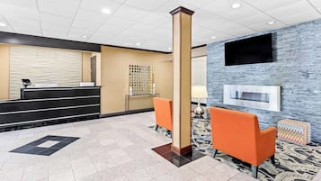 A reception area with a check-in counter, a large pillar, two orange chairs, a patterned rug, and a mounted TV on the wall.