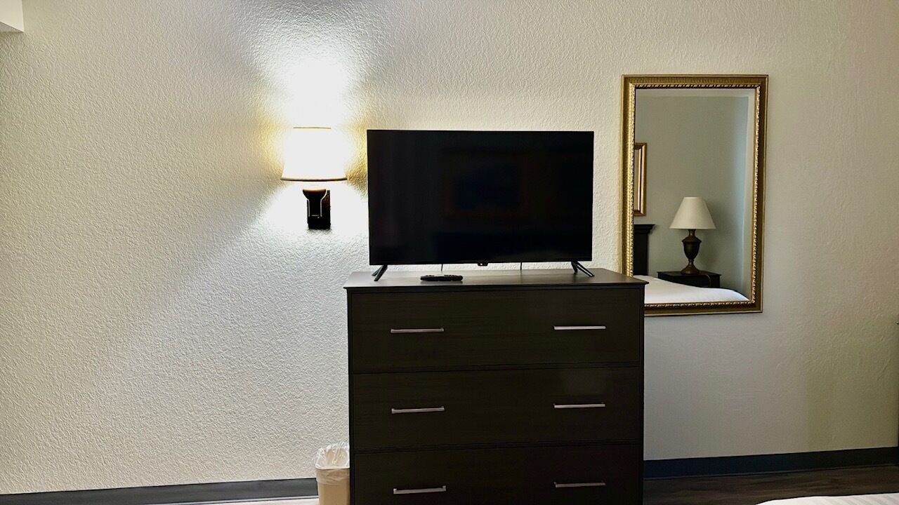 Une chambre d’hôtel équipée d’un téléviseur à écran plat posé sur une commode, d’une lampe fixée au mur et d’un miroir avec un cadre doré.