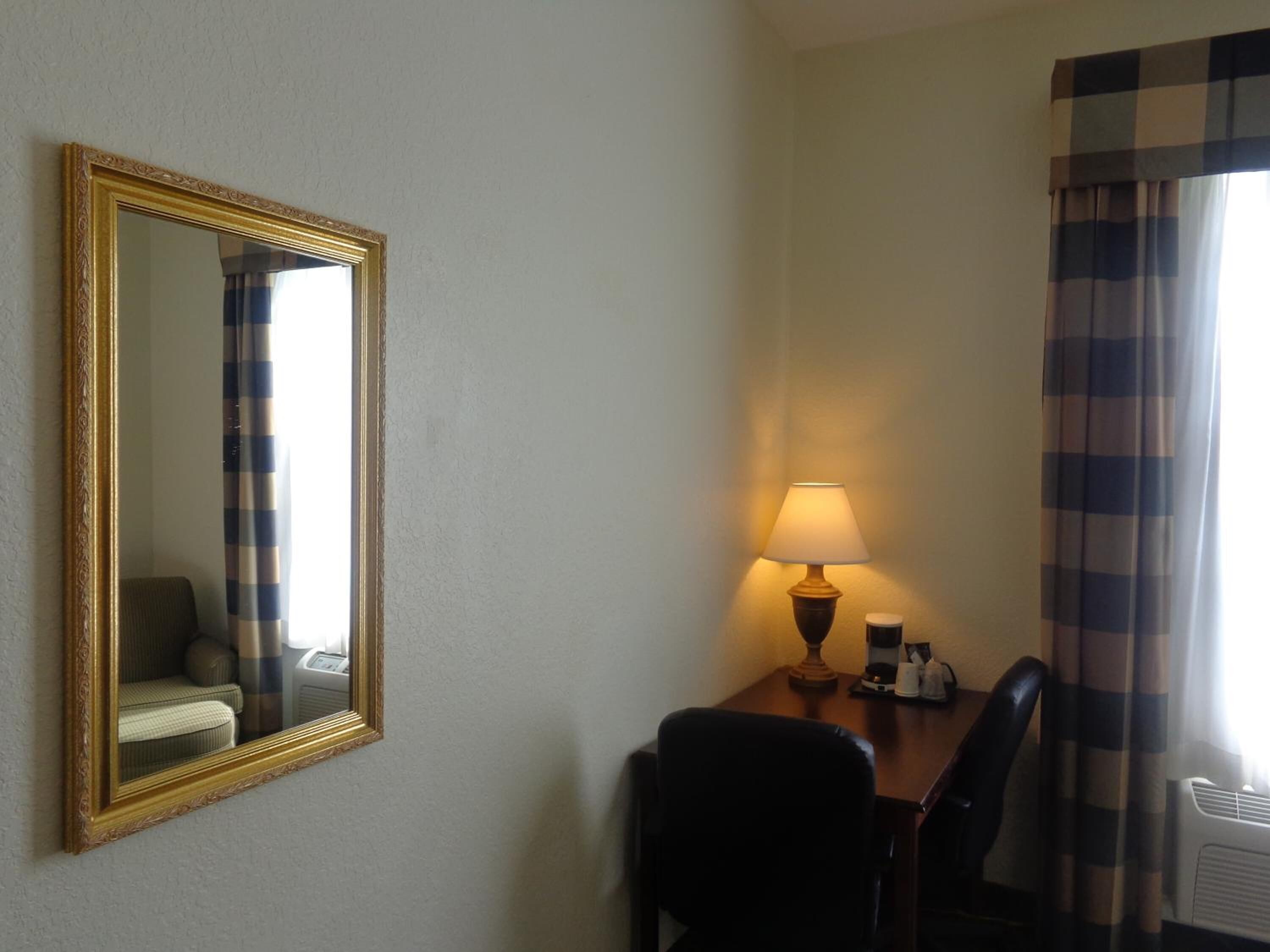 A hotel room with a large mirror, a desk with a lamp, two chairs, and a window with curtains.