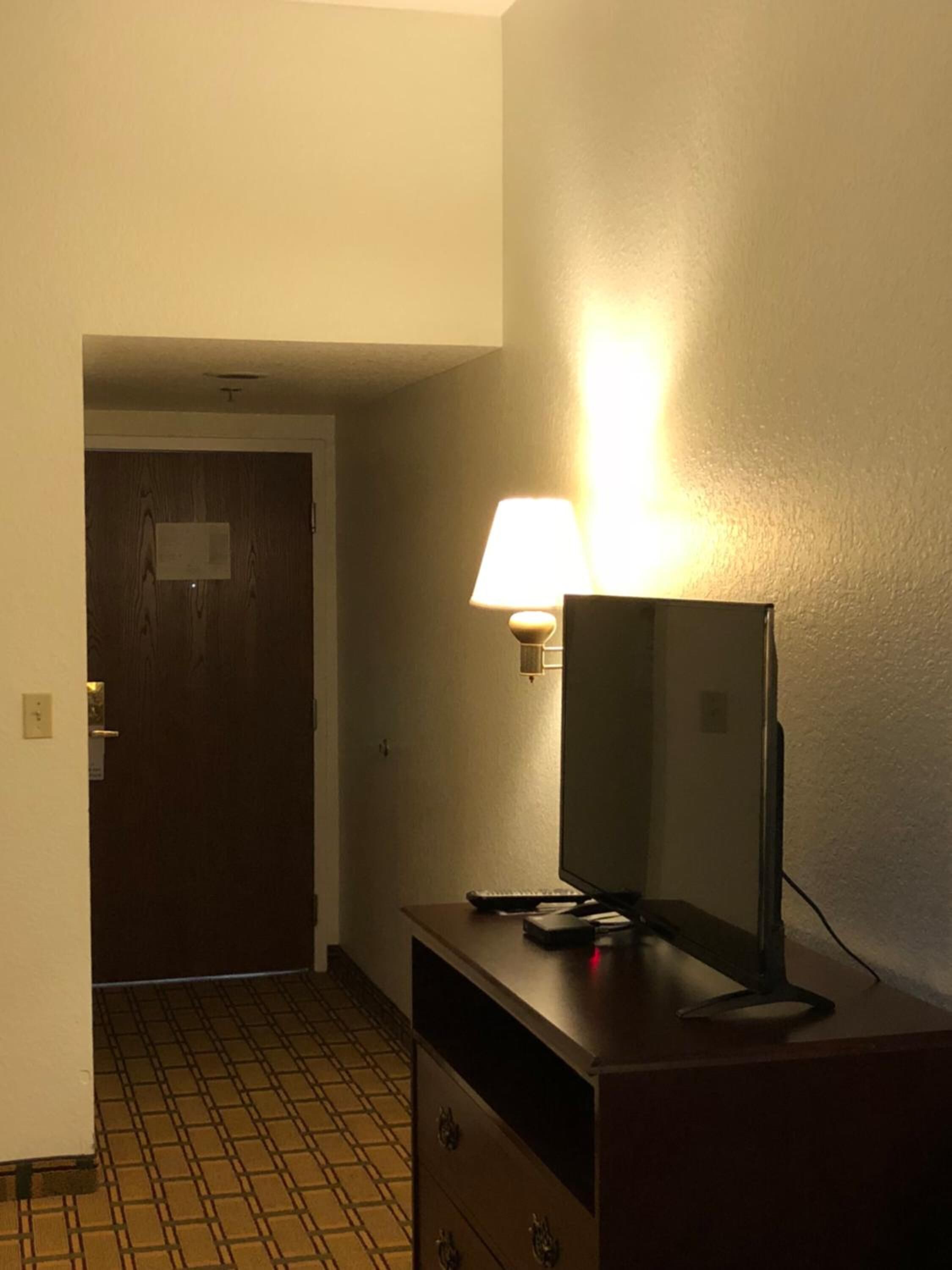 A hotel room with a wooden dresser, a mirror, and a lamp. There is a door leading to another room and a tiled floor.