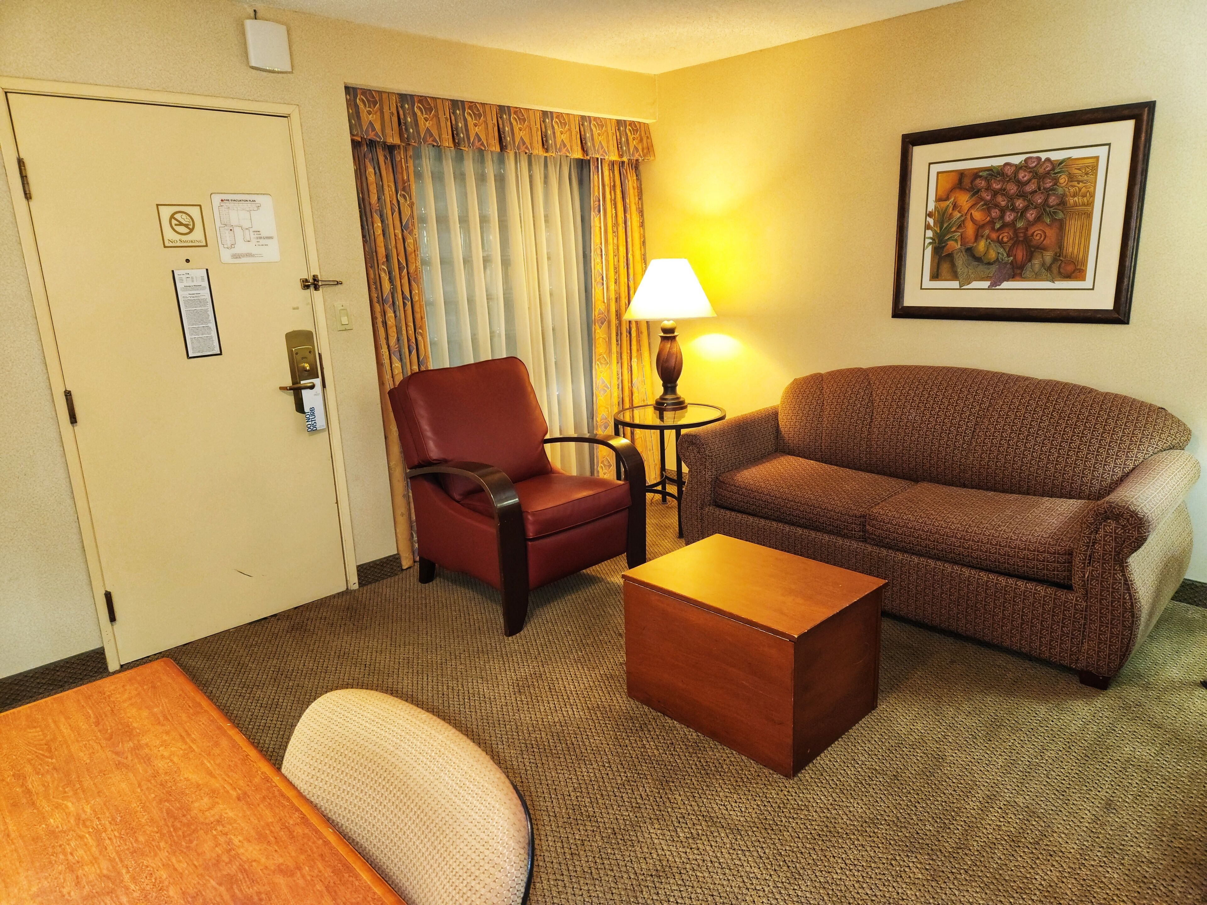 Habitación de hotel con un sillón rojo, un sofá marrón, una mesa de centro de madera y una lámpara. Hay una puerta con un cartel de 'No Fumar' y un cuadro en la pared.