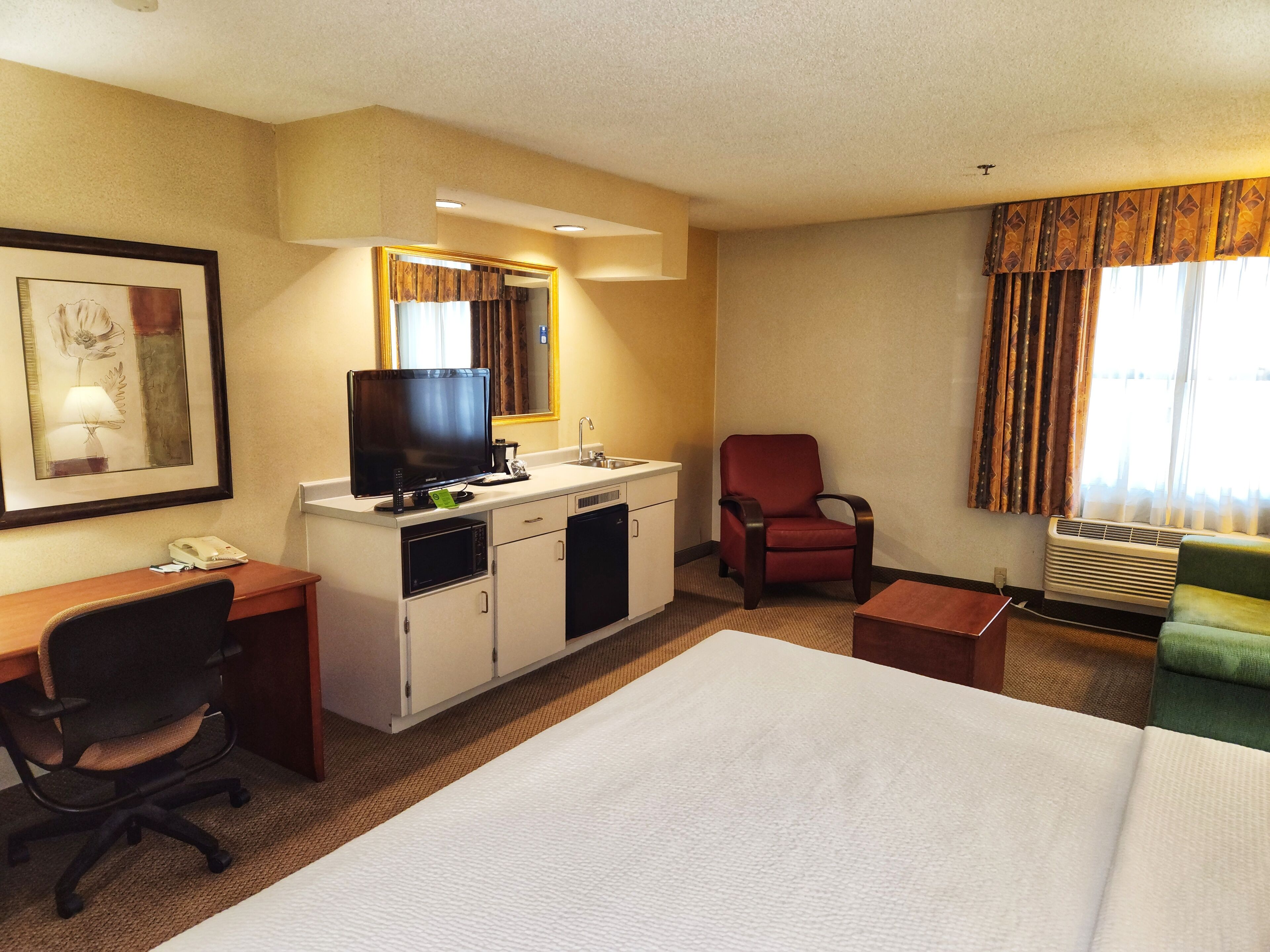 A hotel room with a bed, desk, chair, kitchenette, and a window with curtains.