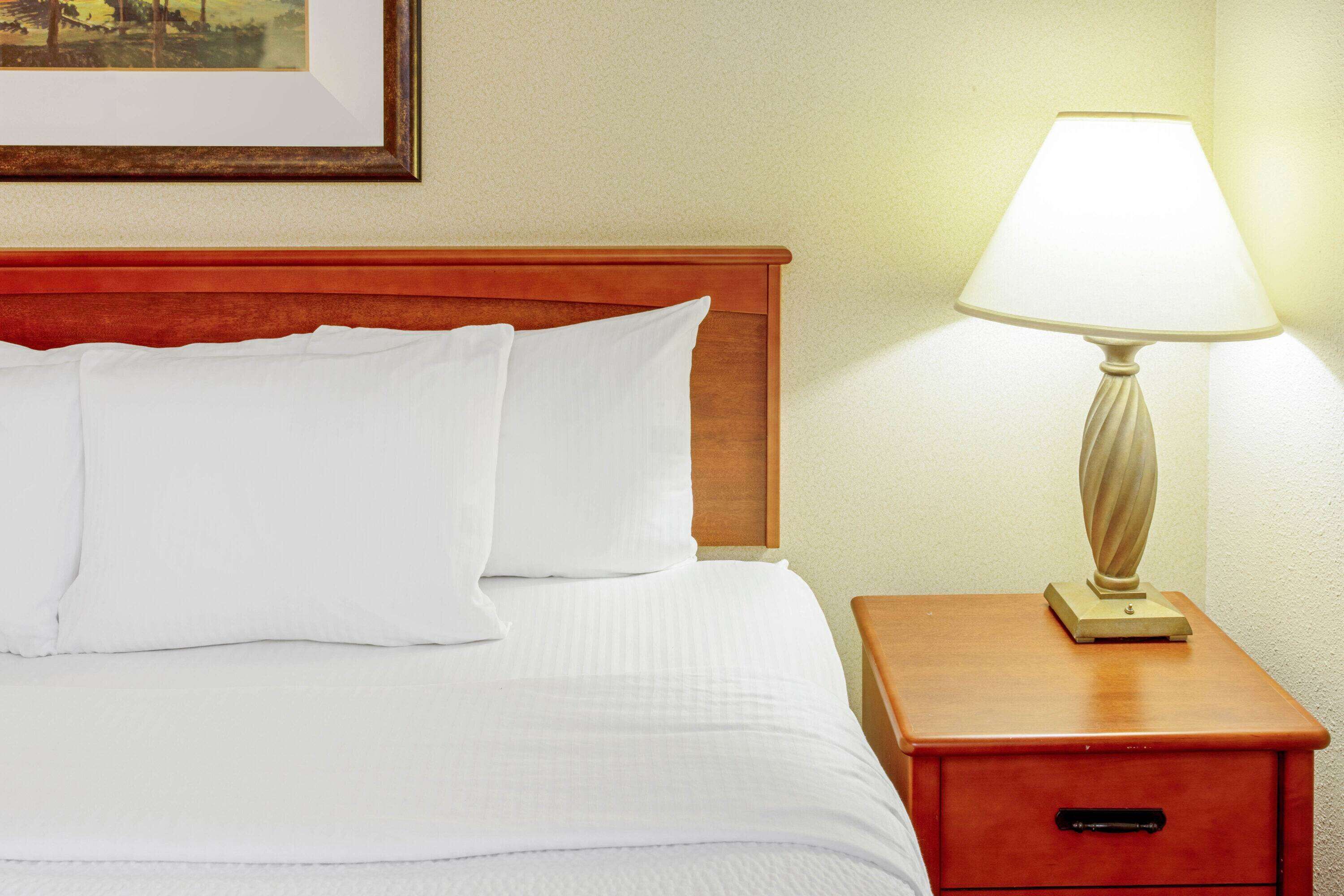A neatly made bed with white linens and pillows, a wooden headboard, and a bedside table with a lamp.