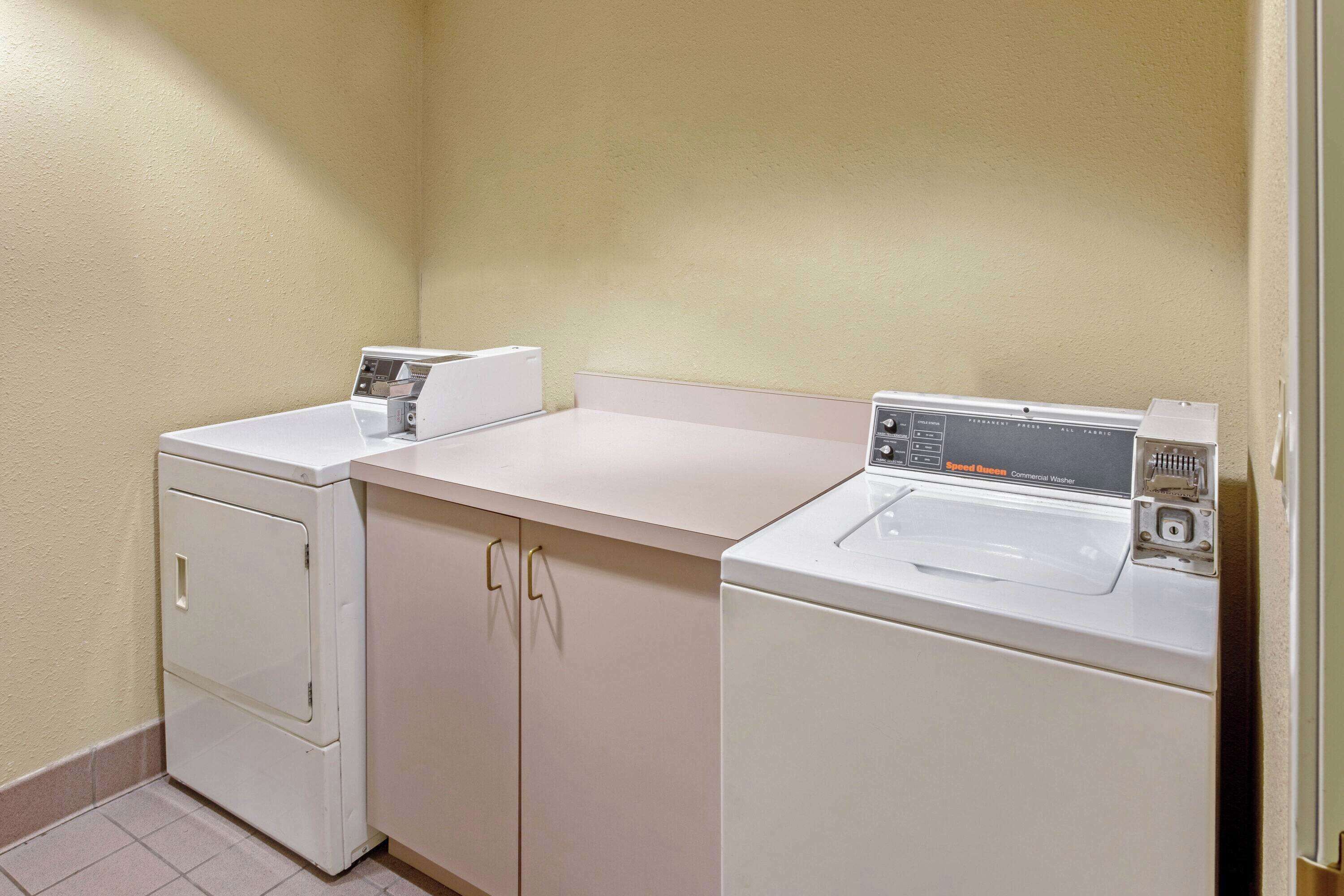 A laundry area with two washing machines and a dryer, a beige wall, and a tiled floor.