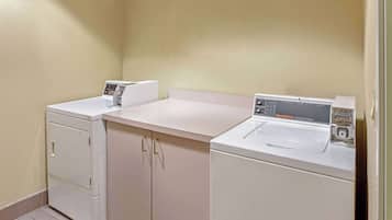 A laundry area with two washing machines and a dryer, a beige wall, and a tiled floor.