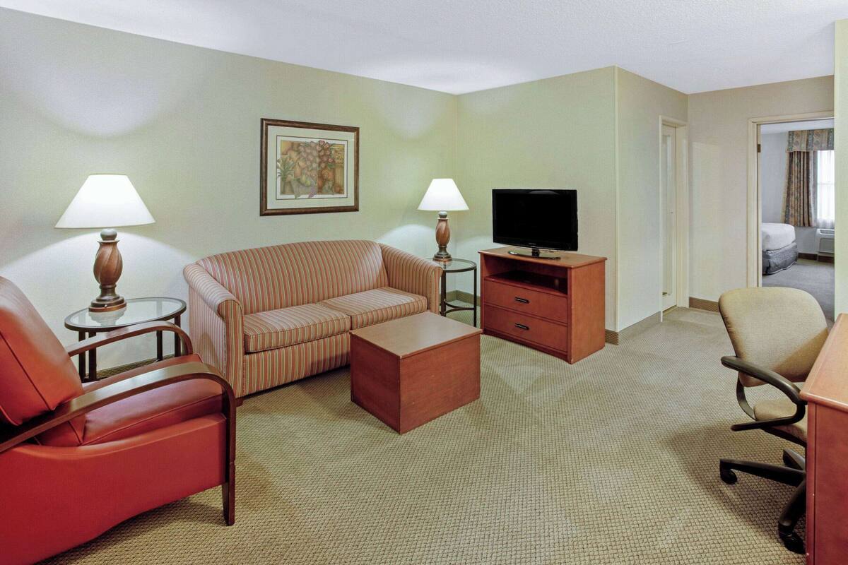 A hotel room with a striped sofa, a red armchair, a wooden coffee table, a television on a wooden stand, and a lamp on a side table.