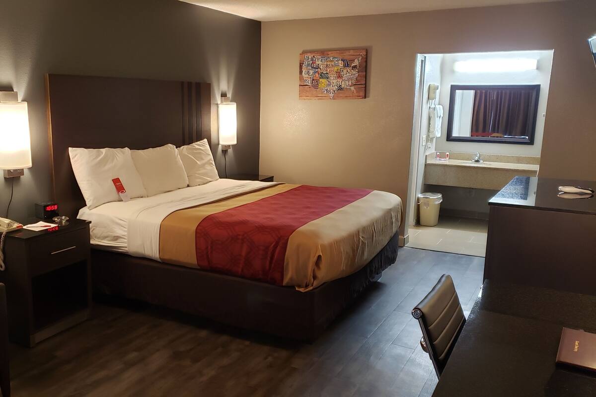 A hotel room with a bed, bedside tables, a desk, a chair, and a bathroom visible through an open door.
