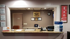 Un mostrador de recepción de hotel con un logotipo enmarcado, un reloj y varios carteles informativos, incluyendo horarios de registro de entrada y salida, desayuno gratuito y un programa de reciclaje.
