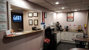 Un vestíbulo de hotel con una recepción, diplomas enmarcados, una máquina de café y un televisor que muestra un noticiero.