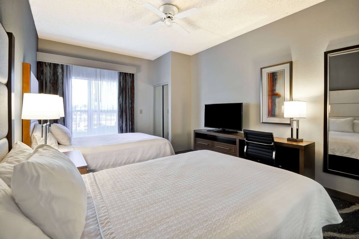 A hotel room with two beds, a television, a desk with a lamp, a ceiling fan, and a large window with curtains.