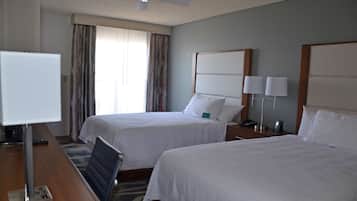 Una habitación de hotel con dos camas, un escritorio con lámpara, un ventilador de techo y una ventana con cortinas.