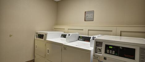 A laundry room with three washers and dryers, a sign indicating laundry products are available for purchase, and a tiled floor.