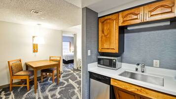 Quarto de hotel compacto com cozinha compacta, área de refeições com mesa e cadeiras, e uma cama visível ao fundo.
