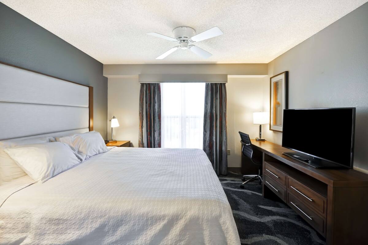 A hotel room with a large bed, a desk with a flat-screen TV, a ceiling fan, and a window with curtains.