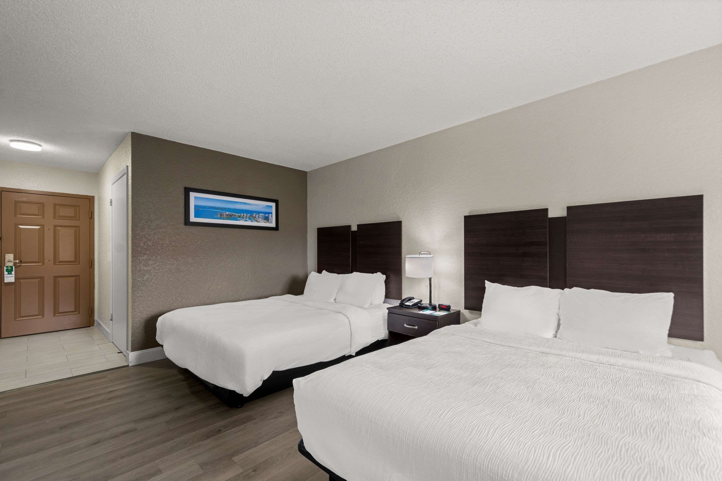A hotel room with two beds, a nightstand with a lamp, a framed picture on the wall, and a door.