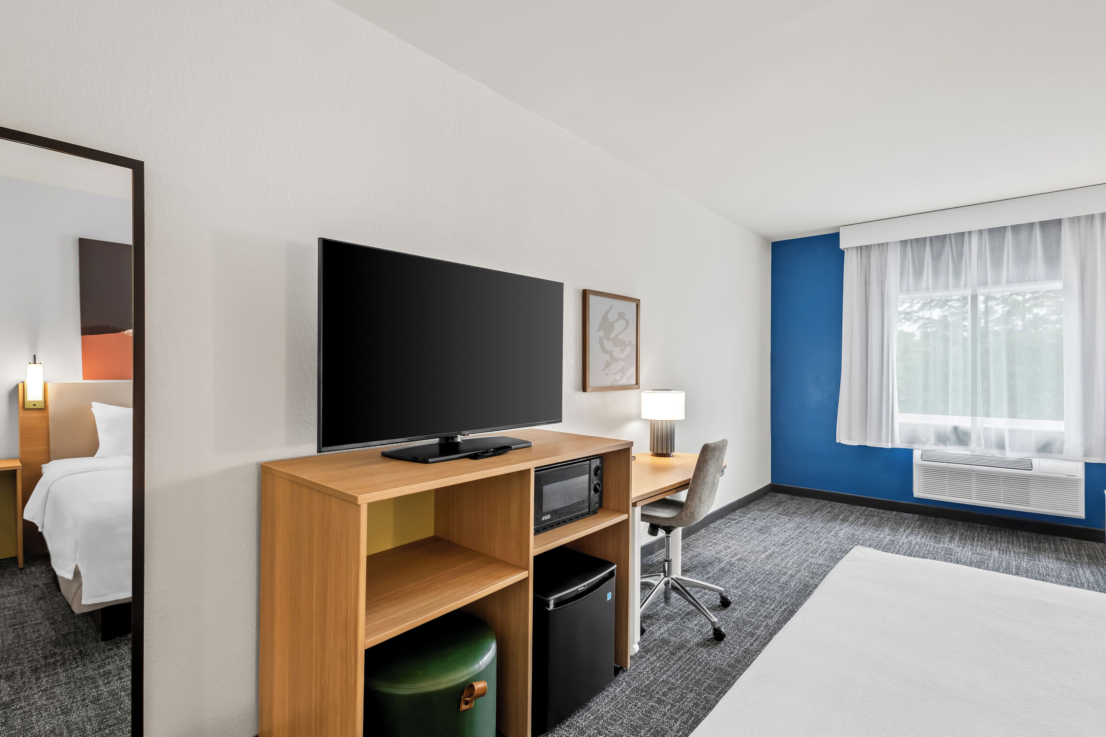 A hotel room with a bed, a desk with a TV, a microwave, a trash bin, and a window with curtains.
