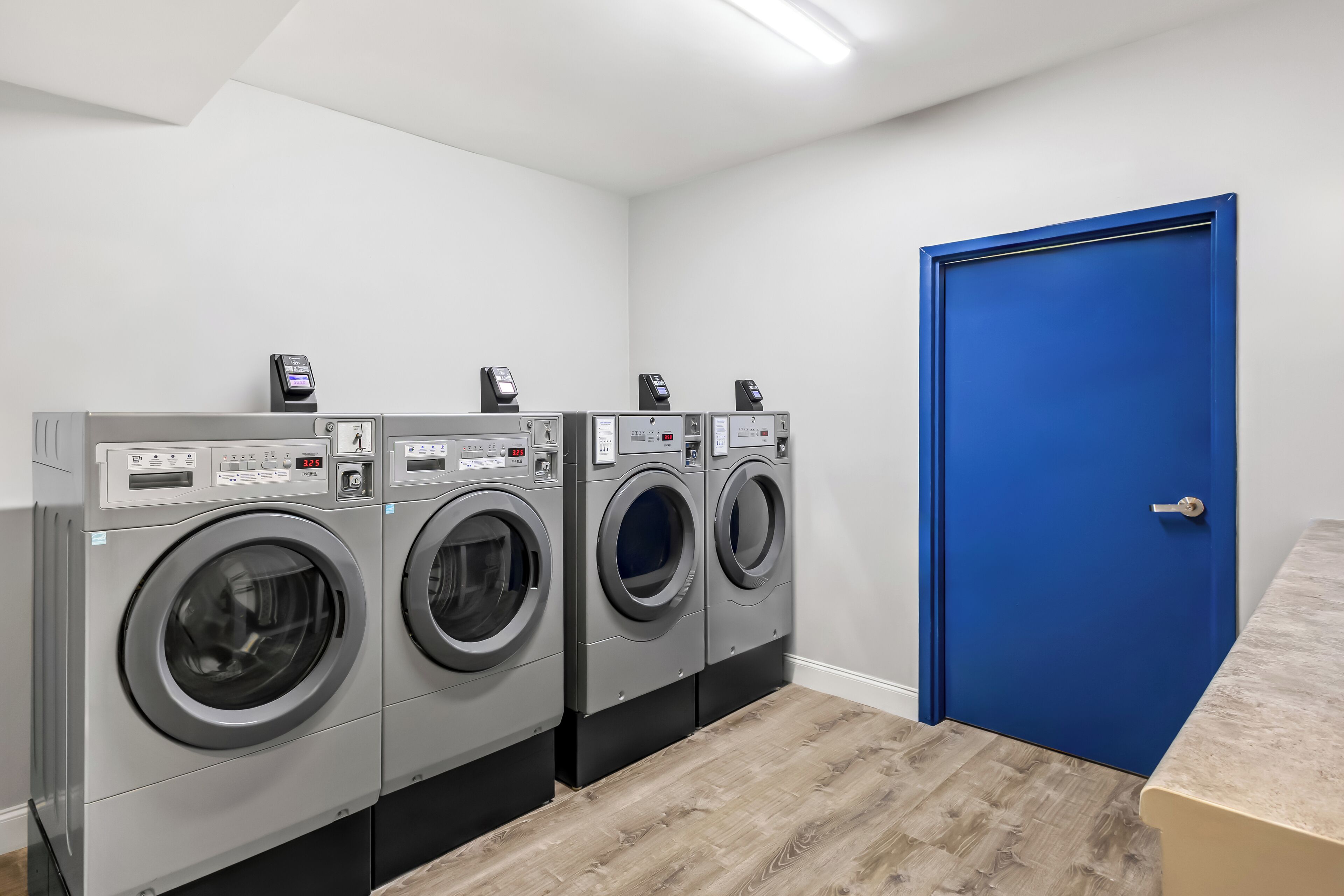 A laundry room with three front-loading washing machines, a blue door, and a wooden floor.